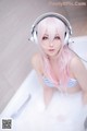 A woman with pink hair sitting in a bathtub with headphones on.