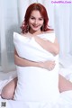 A woman with red hair is sitting on a bed with a pillow.