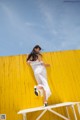 A woman in a white dress leaning against a yellow wall.