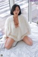 A woman sitting on a bed in a white fur coat.