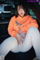 A woman sitting in the back seat of a car wearing white pants and an orange sweatshirt.