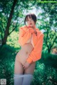 A naked woman in an orange sweatshirt is posing in the woods.