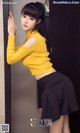 A woman in a yellow top and black skirt leaning against a wall.