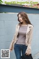 A woman wearing a beige jacket and jeans standing next to a wall.