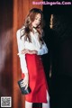 A woman in a white shirt and red skirt leaning against a wooden wall.