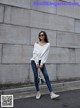 A woman wearing a white sweatshirt and jeans standing in front of a wall.