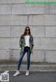 A woman standing in front of a wall wearing a green jacket and jeans.