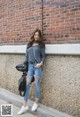 A woman leaning against a brick wall wearing ripped jeans and a gray shirt.