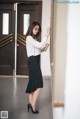 A woman in a white shirt and black skirt leaning against a wall.