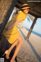 A woman in a yellow dress and hat leaning against a wooden structure.